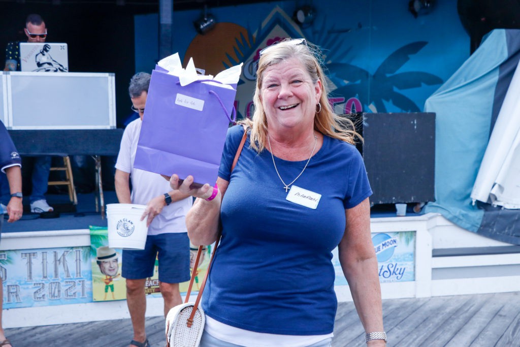 A smiling woman holds a purple gift bag at an outdoor event.