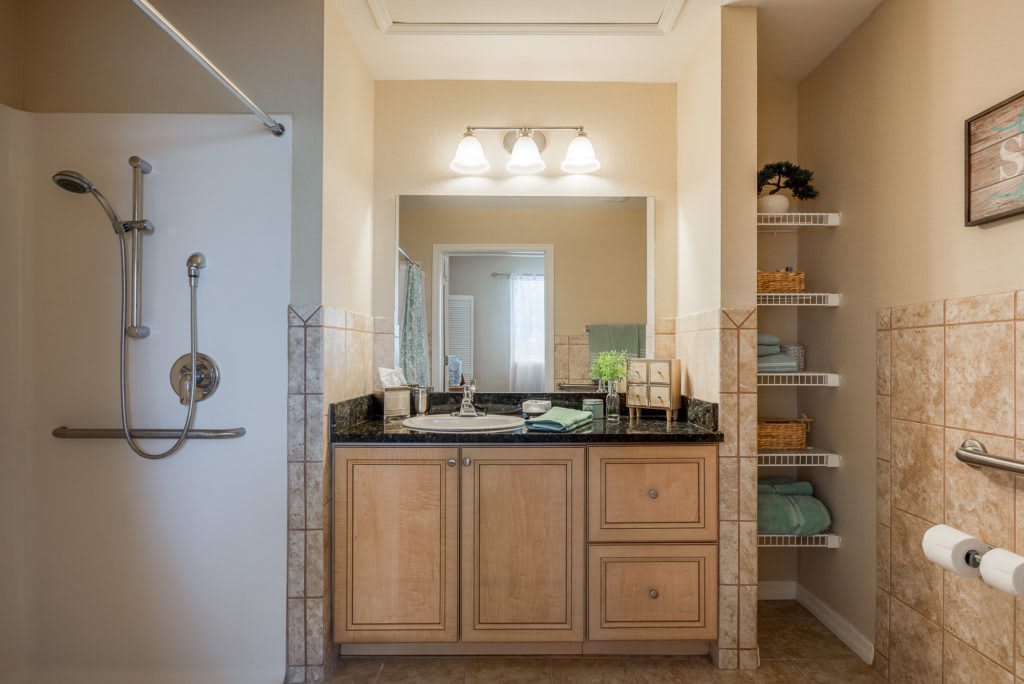 A bathroom featuring a wooden vanity with a large mirror, adjacent to a shower and storage shelves with neatly arranged towels.