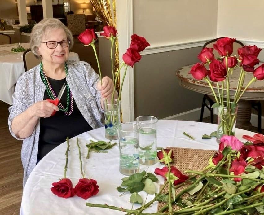 An elderly woman arranges red roses on a table indoors.