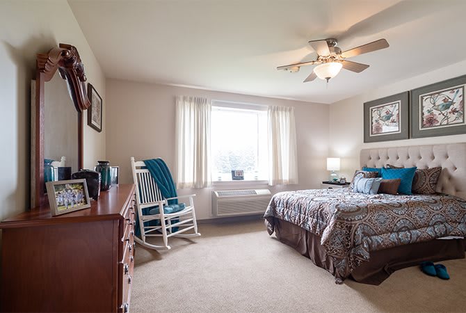 A cozy and well-lit bedroom featuring a bed, rocking chair, and ceiling fan.