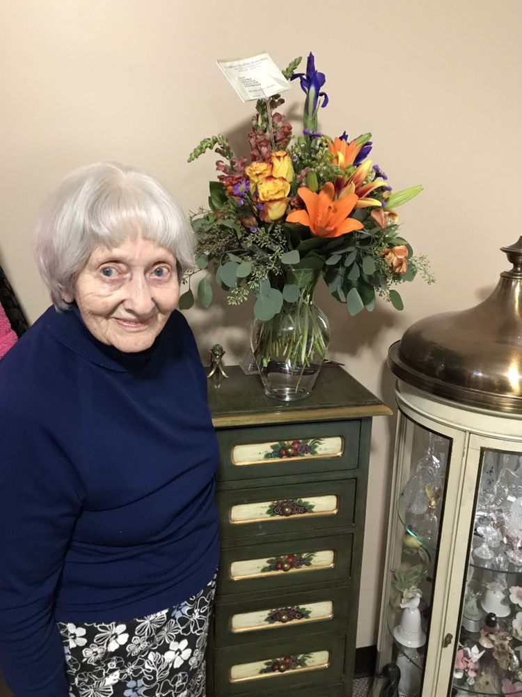 An elderly woman smiling next to a cabinet with a colorful flower arrangement.