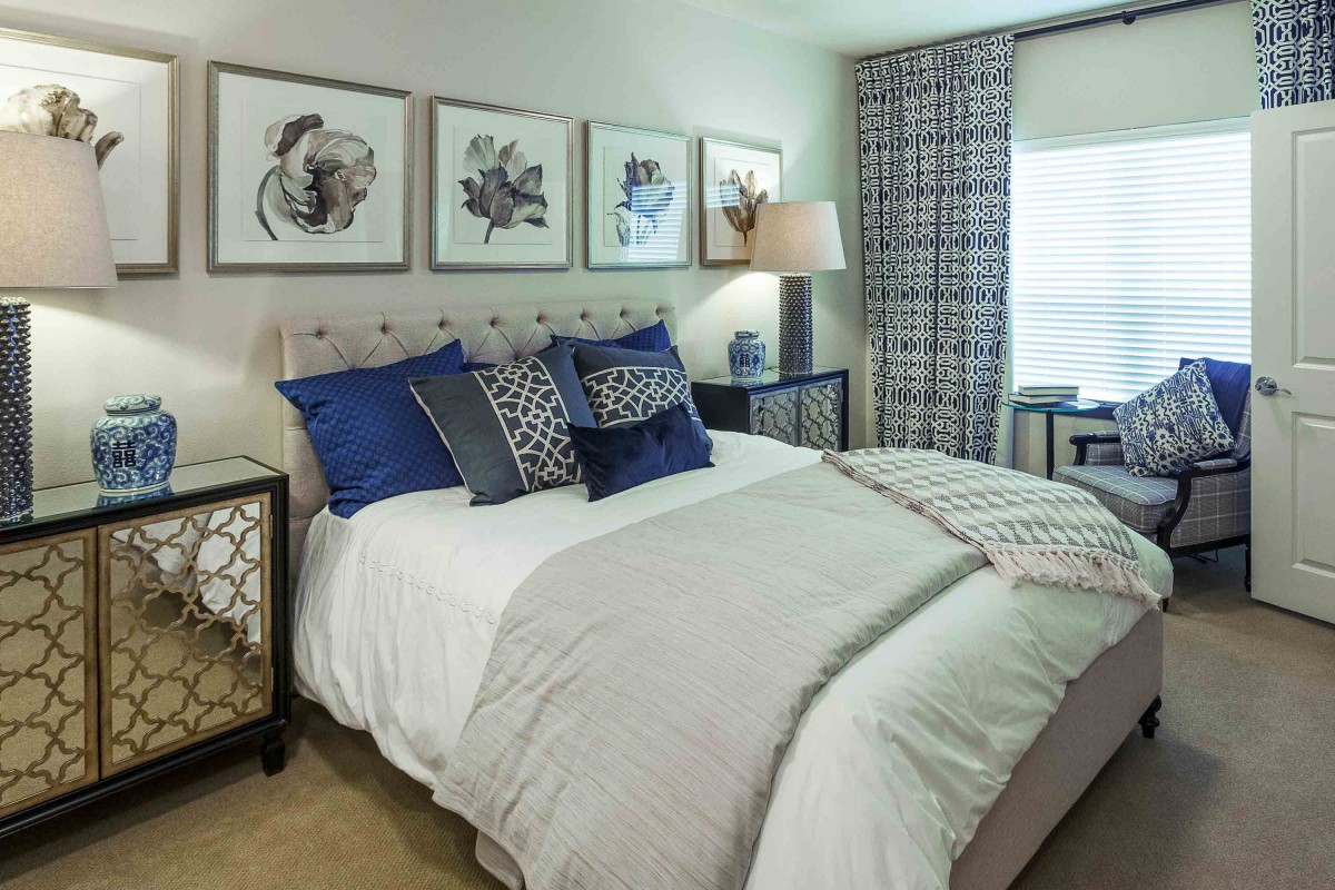A modern bedroom with stylish decor, featuring a bed adorned with blue and patterned pillows, framed artwork on the wall, and elegant nightstands.