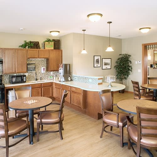 A cozy dining and kitchen area featuring multiple round tables with chairs, wooden cabinetry, and modern lighting fixtures.