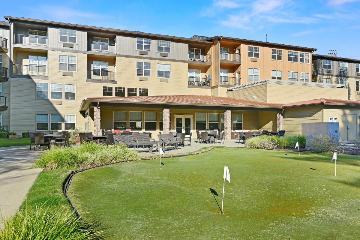 A modern apartment building with a small putting green in the foreground, surrounded by landscaped greenery.