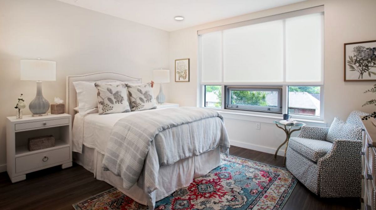 A cozy bedroom with a neatly made bed, colorful rug, and large window letting in natural light.