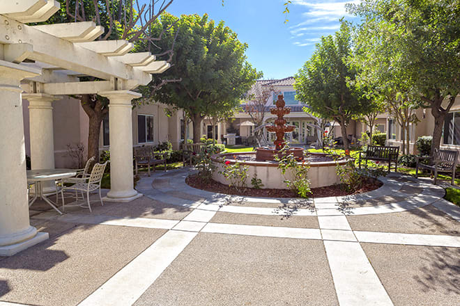 A sunny courtyard with a central fountain, surrounded by trees and outdoor seating under a pergola.