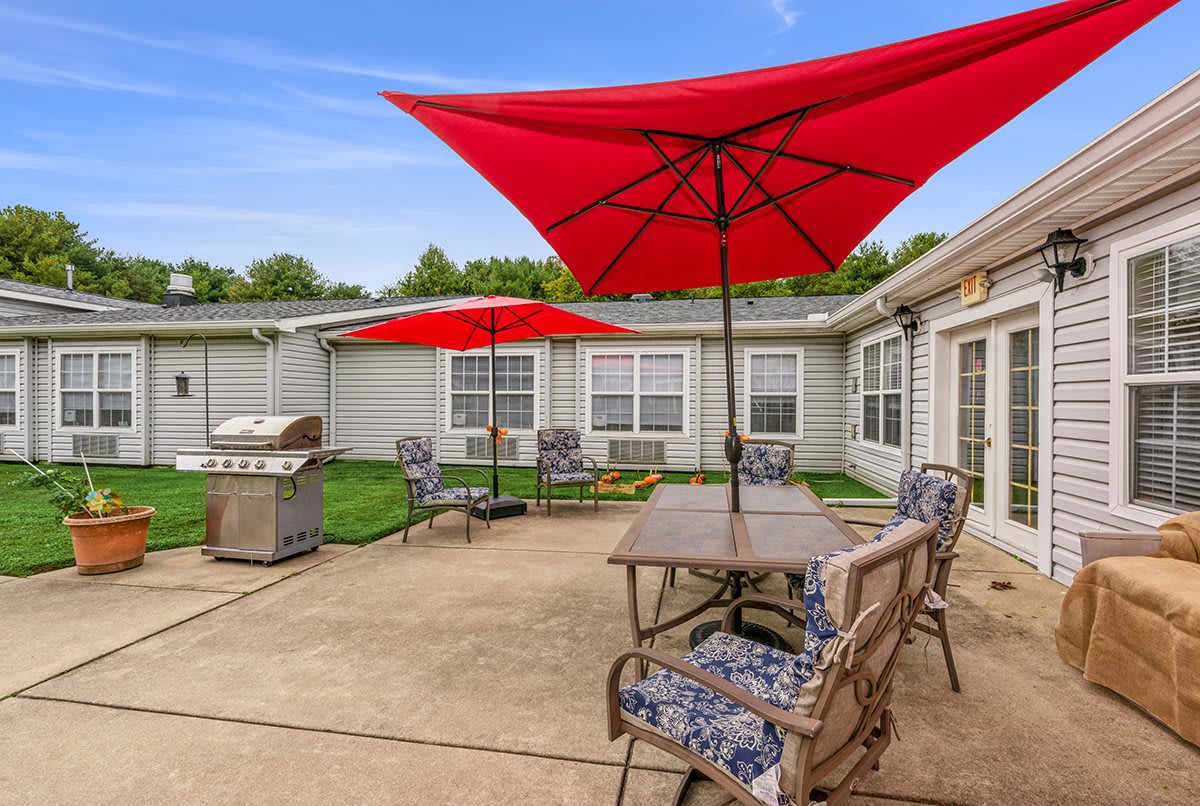 A backyard patio featuring a table, chairs, and red umbrellas, alongside a grill and home exterior.