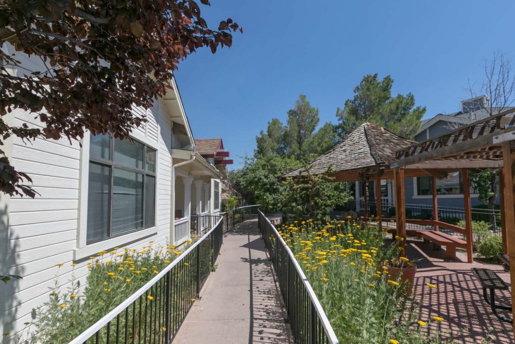 A pathway lined with flowers and greenery leads between houses, featuring a gazebo under a clear blue sky.