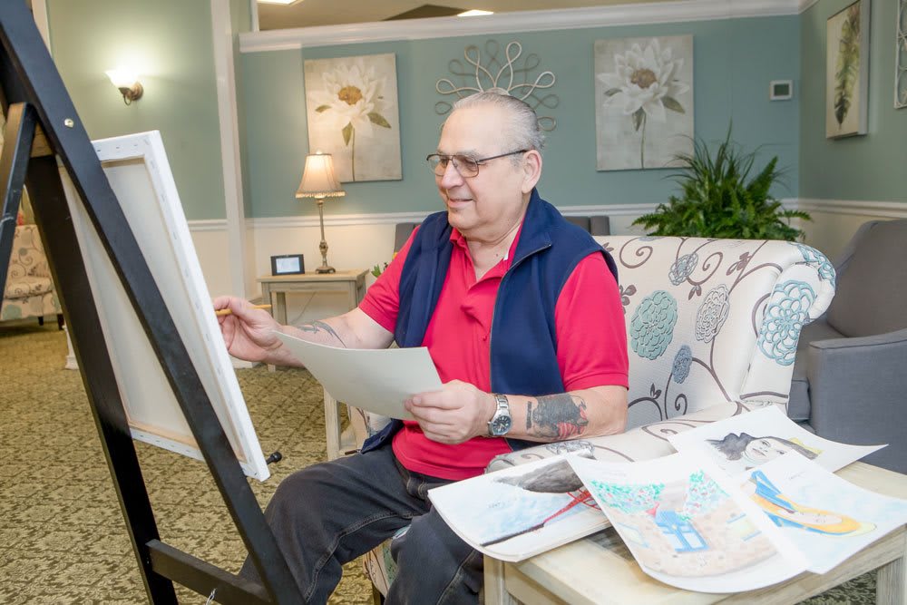 An elderly person enjoys drawing at home, surrounded by newspapers and wearing casual clothes.