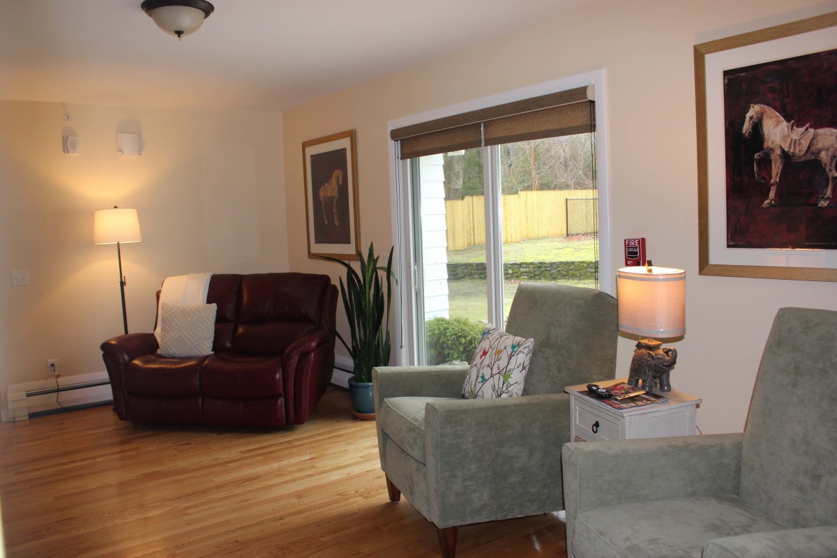A cozy living room with armchairs and a recliner, featuring a lamp on a side table and a large window overlooking the yard.