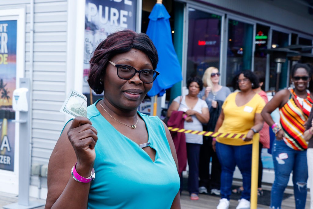 A woman holding money and smiling outdoors, with a crowd in the background.