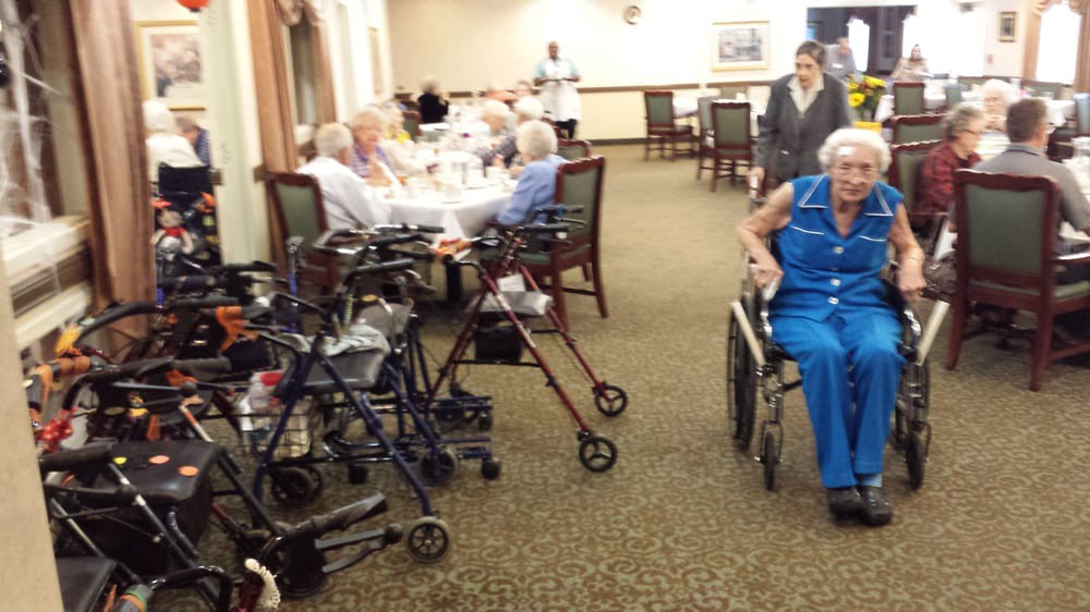 Residents in a senior living facility dining room, with wheelchairs and walkers nearby.