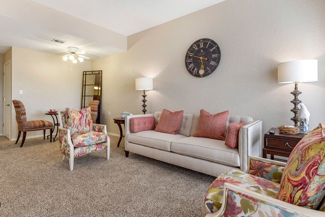 A cozy living room featuring a sofa with pink cushions, floral armchairs, and a large wall clock.