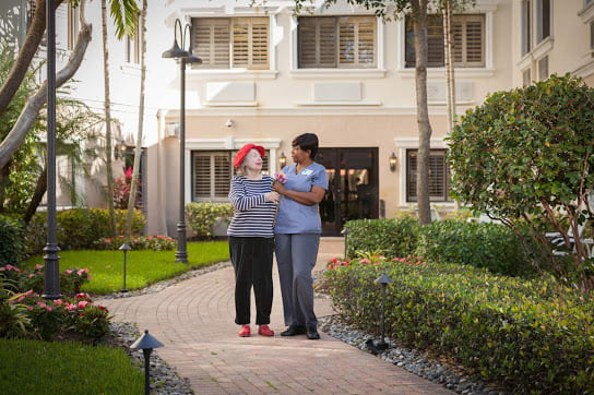 A caregiver assisting an elderly person on a walk in the garden of a healthcare facility.