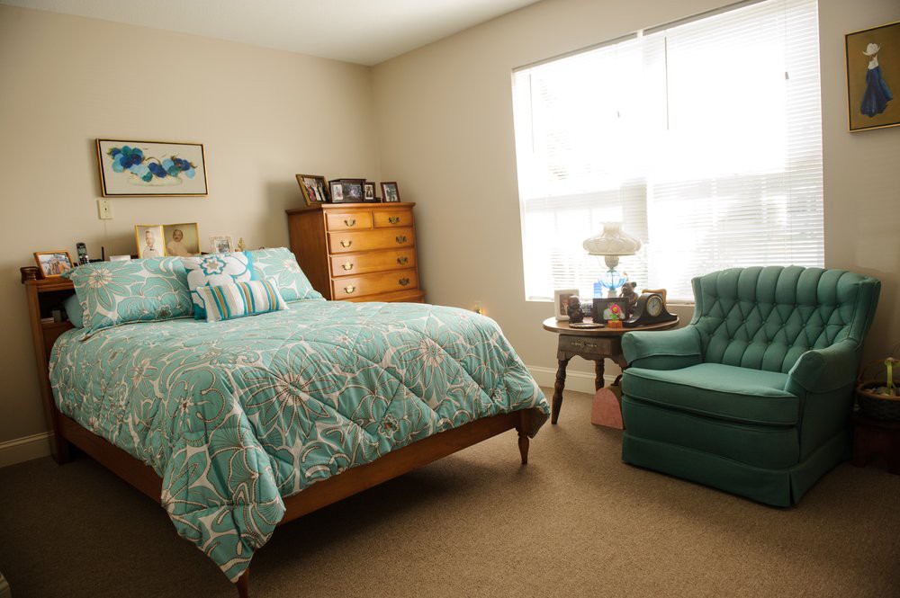 A cozy bedroom with teal bedding, an armchair, and a wooden dresser beside a large window.