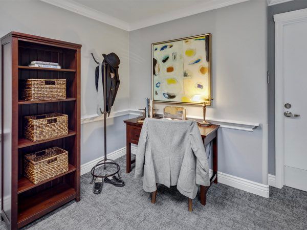 A small office corner with a desk, chair, and a suit jacket draped over the chair. The space includes a shelf with baskets, a coat rack, and a colorful painting on the wall.