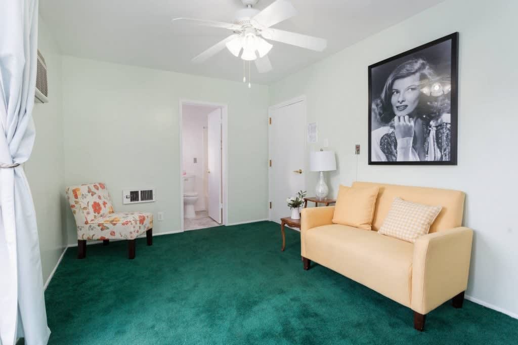 A cozy living room with a green carpet, featuring a beige sofa and a floral armchair. A ceiling fan hangs overhead, and there's a black and white portrait on the wall.