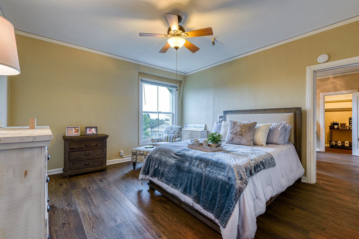 A cozy bedroom with a large bed, wooden flooring, and a ceiling fan.