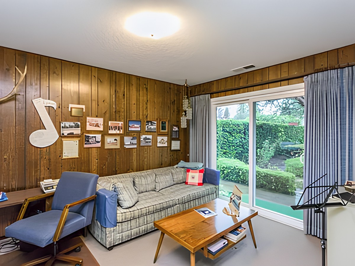 A cozy mid-century modern living room with wood paneling, featuring a sofa, coffee table, armchair, and sliding glass doors opening to a garden view.