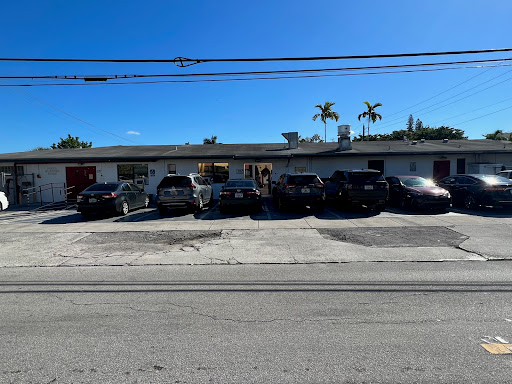 A sunny day shows a parking lot filled with cars in front of a building surrounded by palm trees.