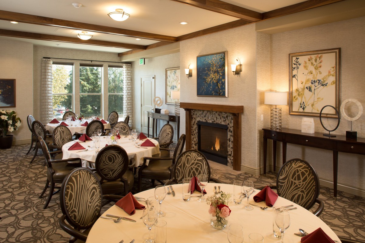An elegant dining room with round tables set for a meal, featuring a cozy fireplace and tasteful artwork on the walls.