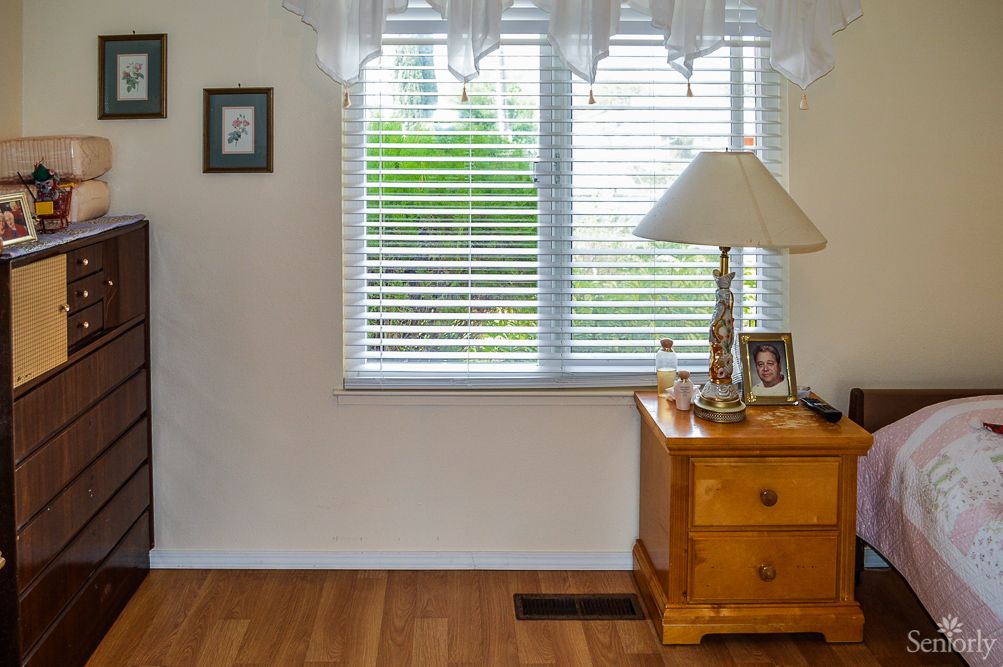 A cozy bedroom corner with a wooden nightstand, lamp, and window with blinds.