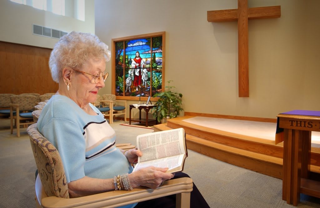 An elderly woman reads a Bible in a church with stained glass and a cross in the background.