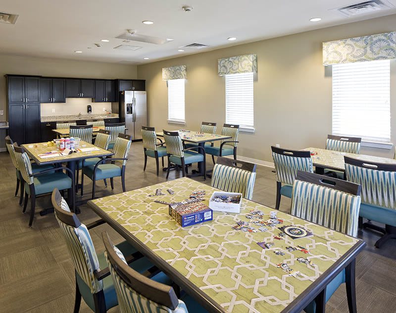 A spacious community dining and activity area with tables, chairs, and a kitchen in the background.