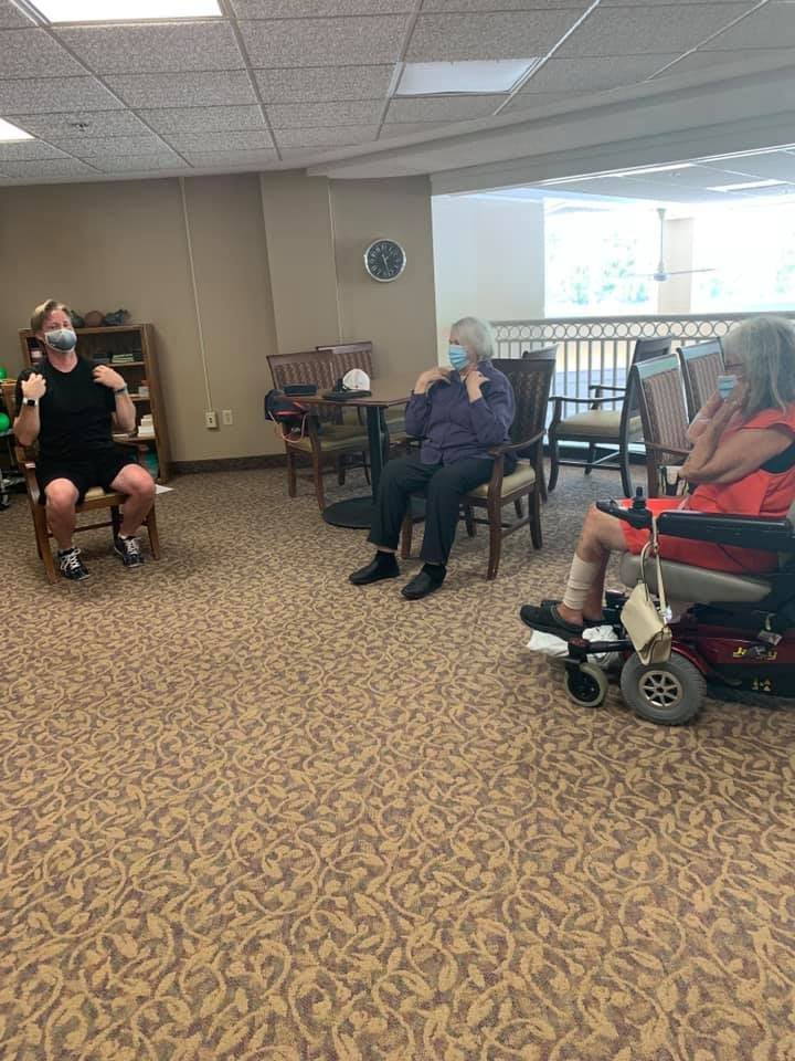 A group of seniors sitting in a common area, some wearing masks and one in a wheelchair.