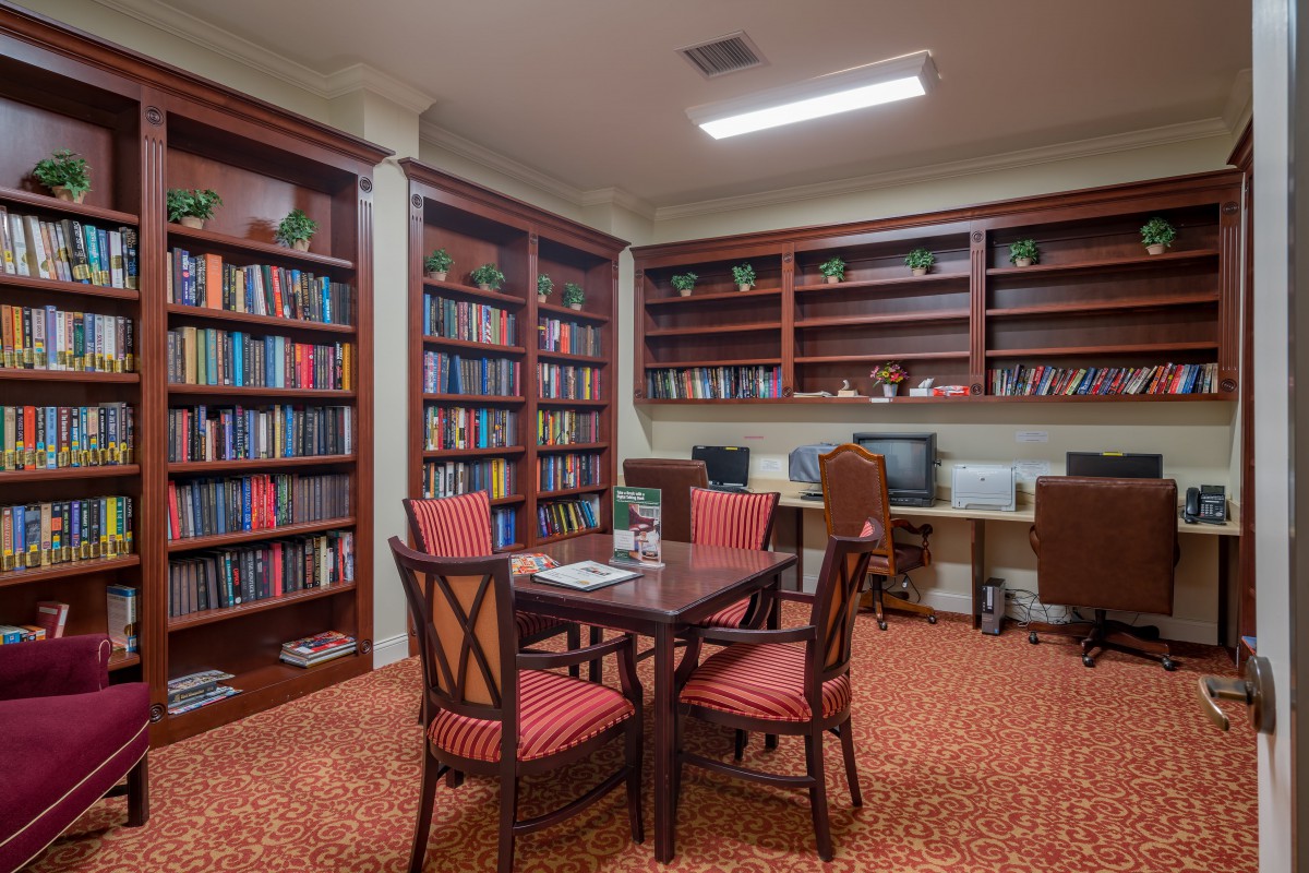 A cozy library room with bookshelves, a table, chairs, and computers.