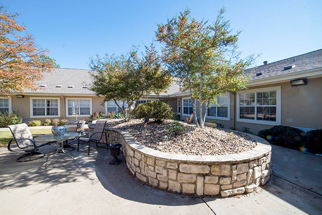 A serene courtyard featuring outdoor seating, surrounded by trees and a stone wall under a clear blue sky.