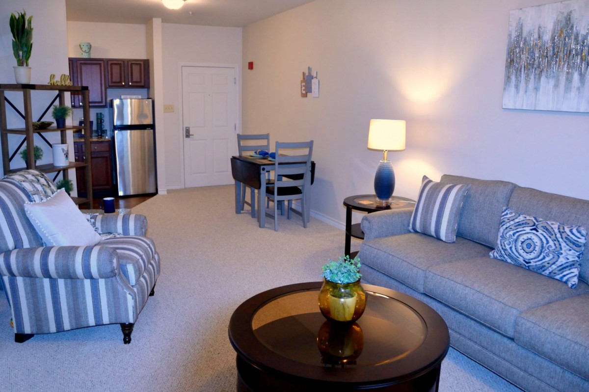 A cozy living room with modern furniture including a sofa, lamp, and coffee table, adjacent to a small dining area and kitchen.