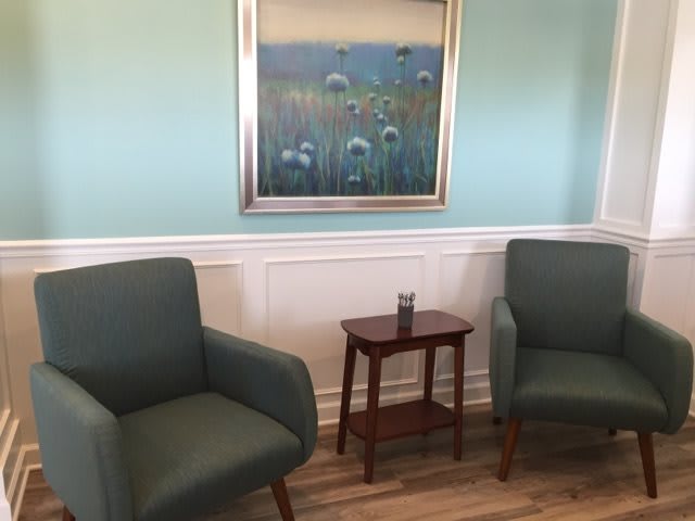 A cozy waiting room with two green chairs and a wooden table beneath a floral painting.