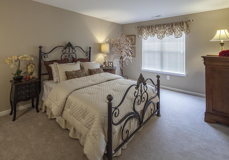 A cozy bedroom with an iron bed frame, soft lighting from a lamp, and a window with blinds and drapes.