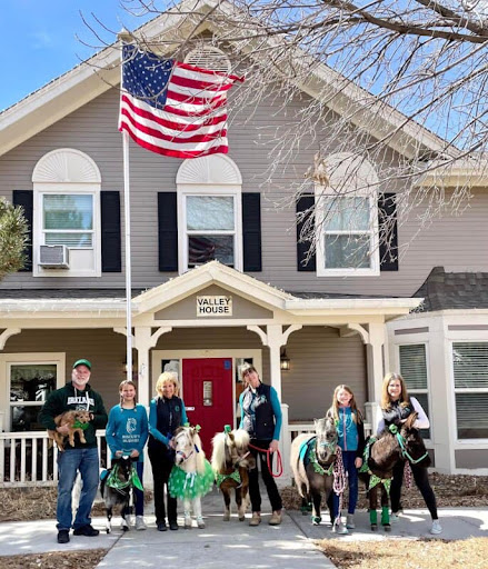 A festive gathering in front of a charming house, featuring a group of people with horses and an American flag waving proudly.