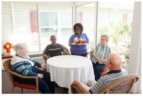 A caregiver serves drinks to a group of seniors gathered around a table in an indoor setting.