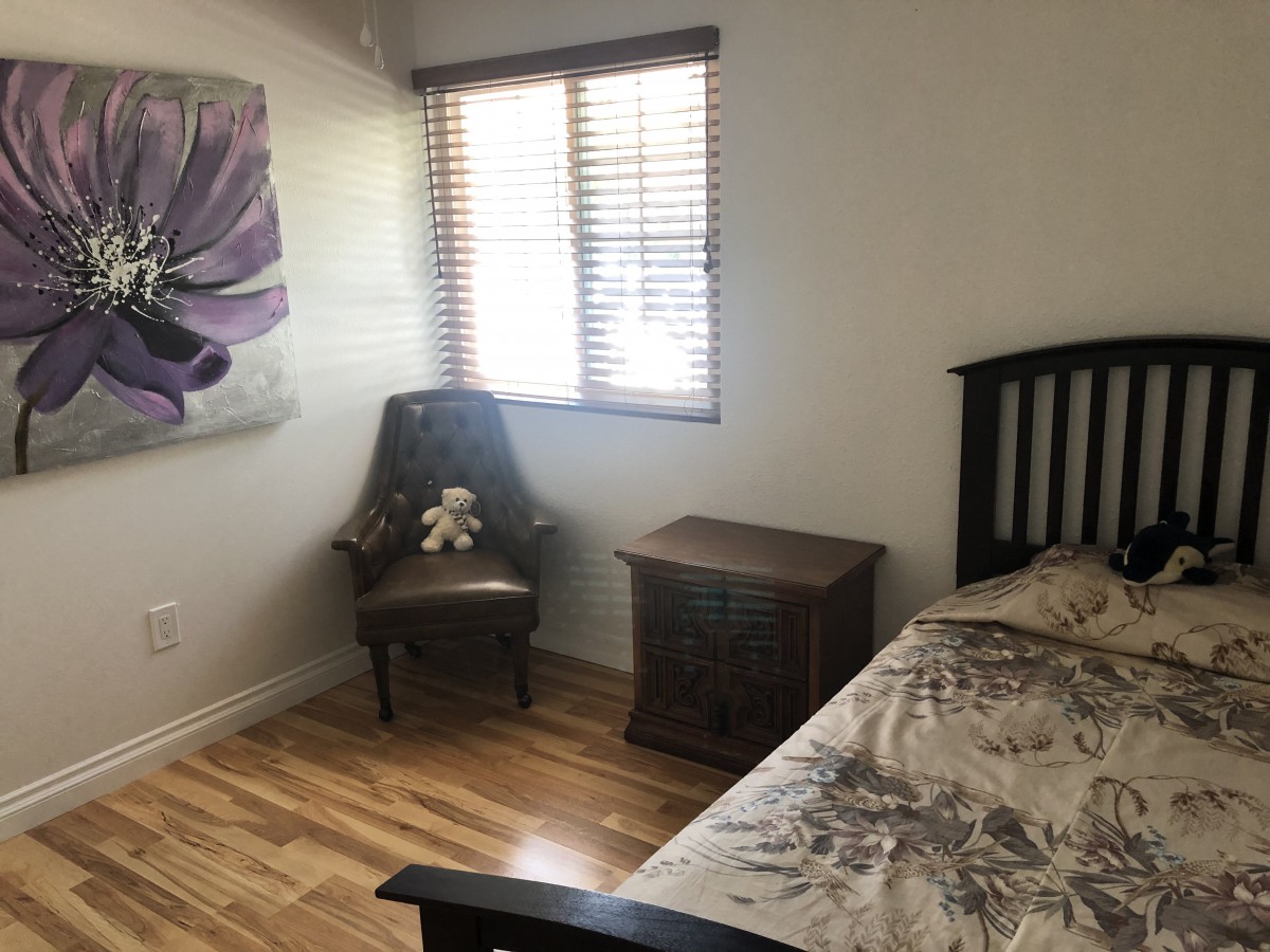A small bedroom with a bed, chair, nightstand, and a flower painting on the wall.