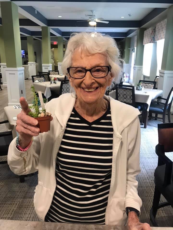 An elderly woman with glasses smiling and holding a small potted plant indoors.
