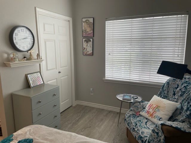 A cozy bedroom with muted colors, featuring a patterned chair, side table, dresser, and window blinds.