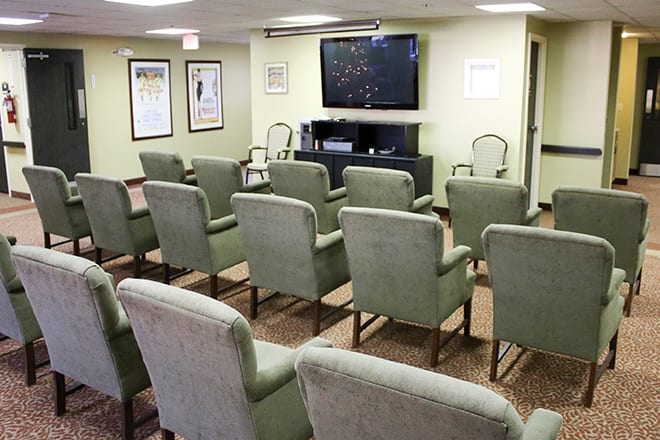 An empty room with arranged chairs facing a large television screen, resembling a waiting or conference area.