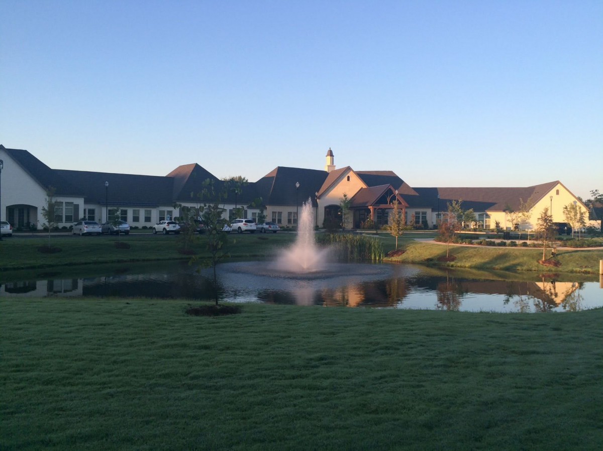 A scenic view of a building with a fountain in the foreground, reflecting in a tranquil lake at sunset.