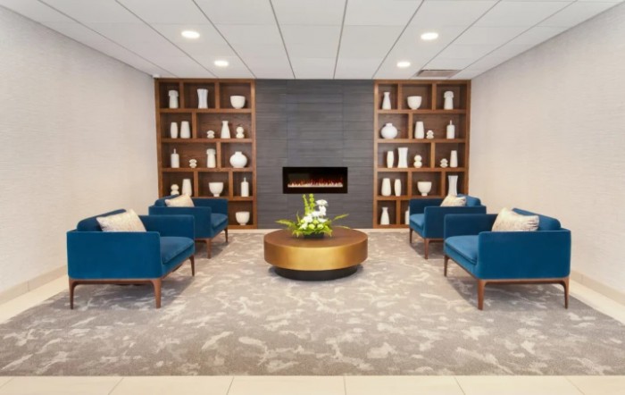 A modern lobby with blue seating, a central round table, and a stylish fireplace surrounded by decorative shelves.