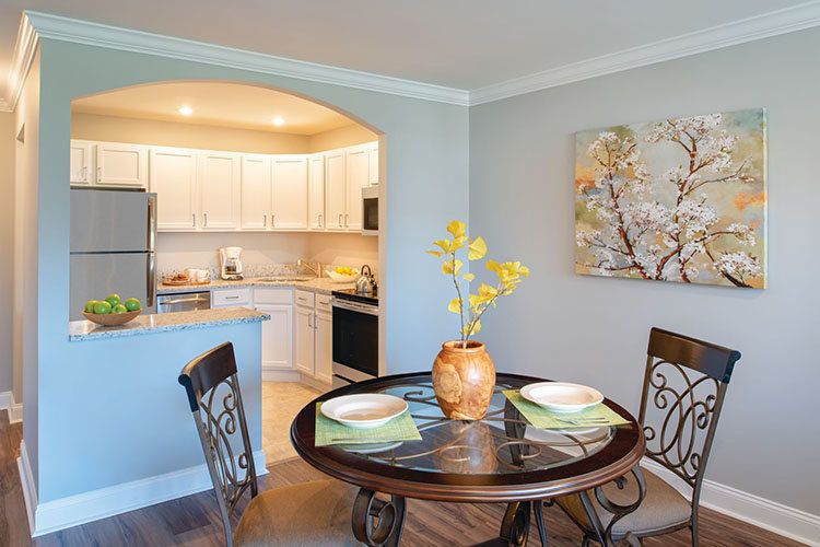A modern dining area with a round table set for two, adjacent to an open kitchen and decorated with a potted plant and artwork on the wall.