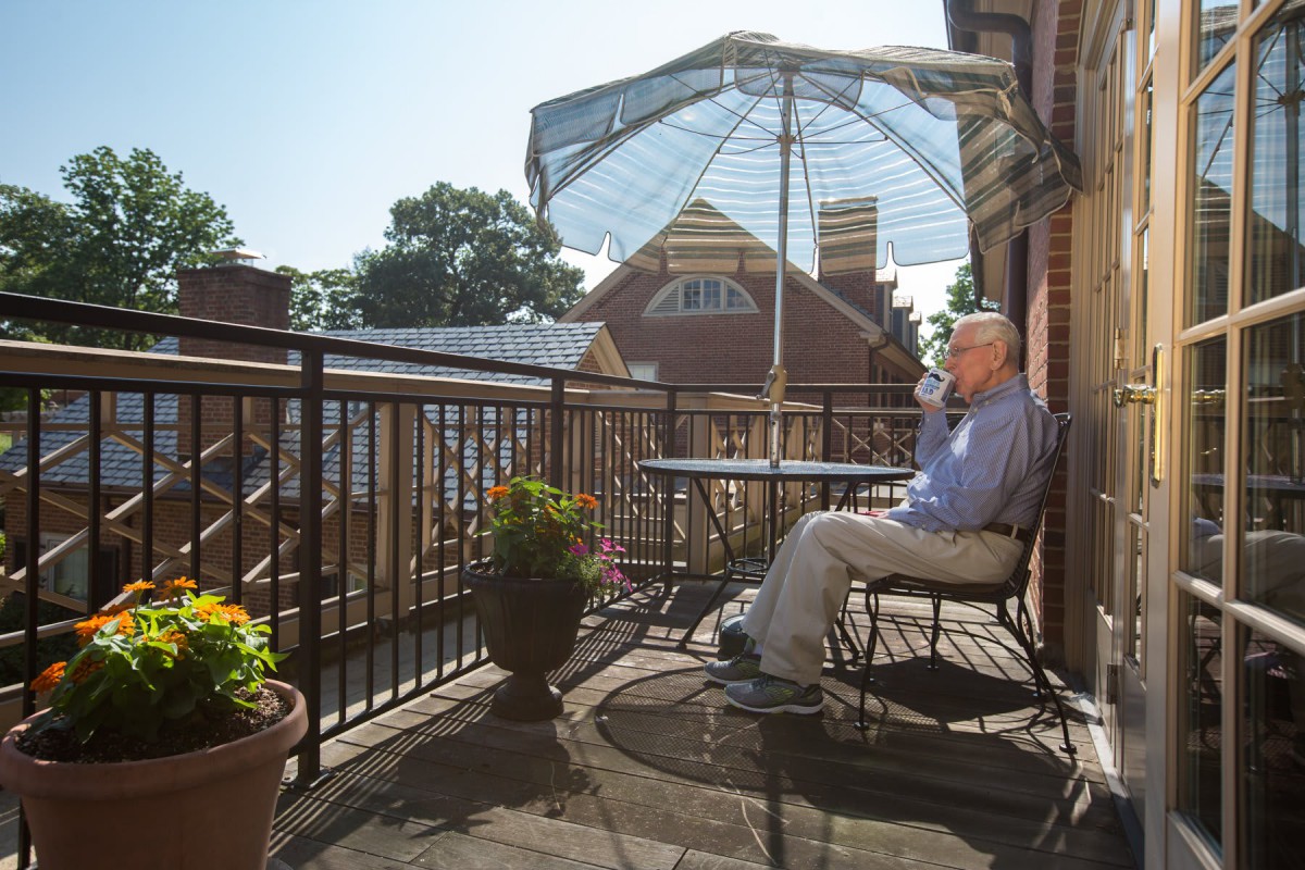 A man enjoys a sunny day on a balcony, sitting under an umbrella with potted plants nearby.