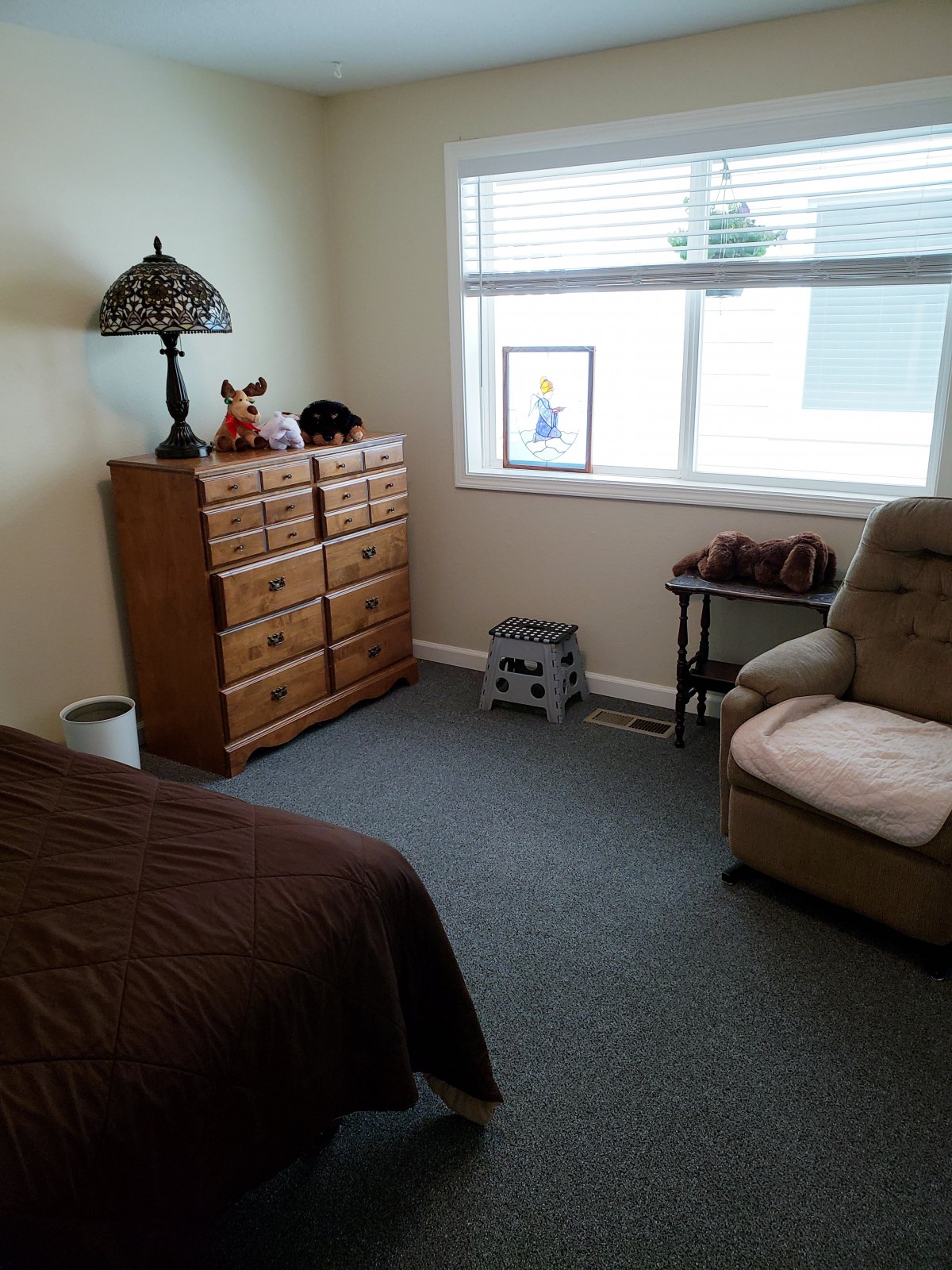 A cozy bedroom with a dresser, armchair, and a bed, illuminated by natural light from the window.