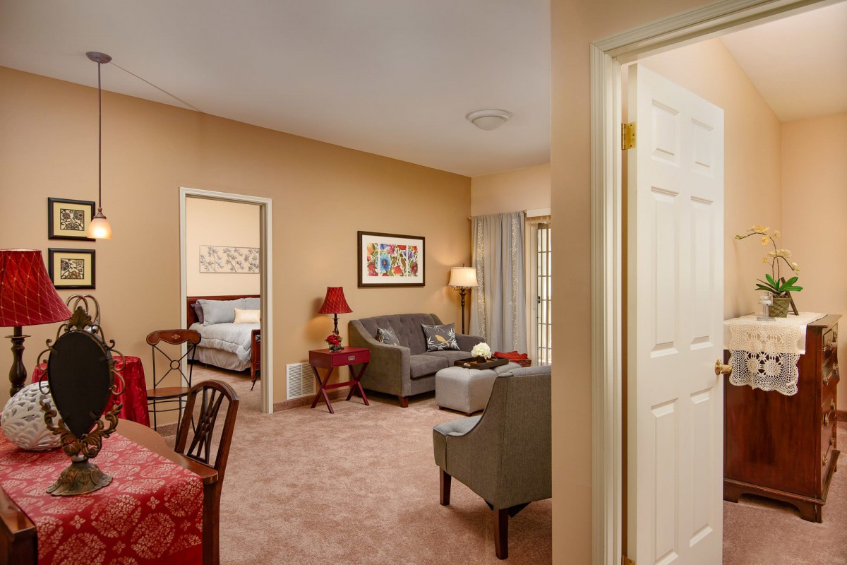A cozy living room with a sofa, armchair, and table, leading into a bedroom.