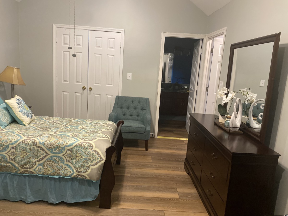 A cozy bedroom featuring a bed, chair, dresser with mirror, and an open door leading to a bathroom.