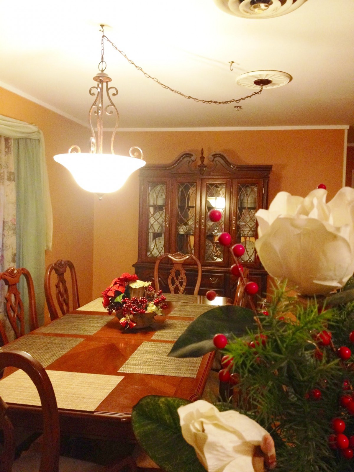 A warmly lit dining room with a large wooden table, ornate chandelier, and decorative floral arrangements.