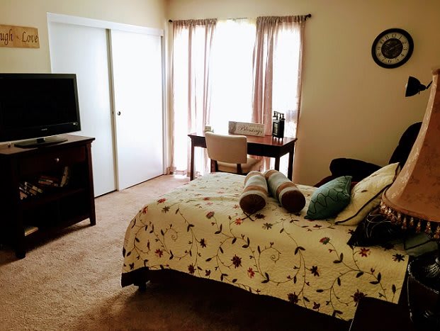A cozy bedroom featuring a neatly made bed, a television, and a desk near the window with curtains.