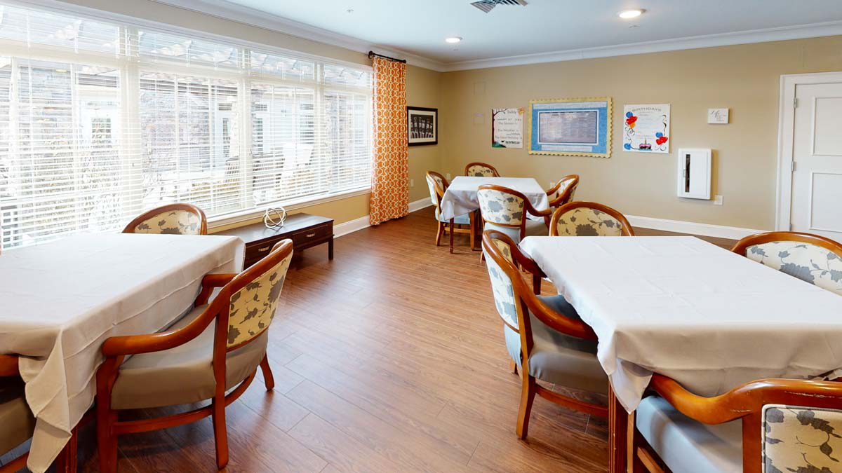 A cozy dining area with several tables and chairs, featuring large windows and wooden flooring.